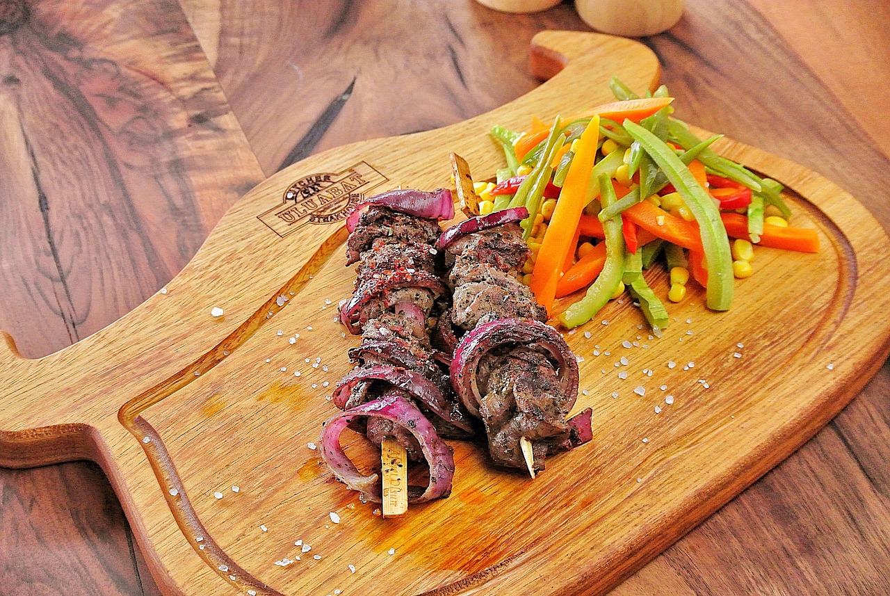 나무 도마 위에 붉은 양파와 함께 구운 고기 꼬치와 채소 볶음이 담겨 있습니다.